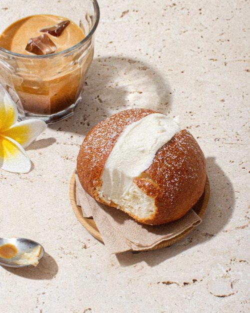 A brioche bun filled with ice cream, beside a glass of coffee. Decorated with a frangipani flower and spoon